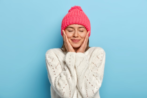 Retrato de uma menina adorável com expressão facial satisfeita tocando ambas as bochechas, mantém os olhos fechados, usa um chapéu rosa e um suéter branco, gosta de poses de inverno contra uma parede azul