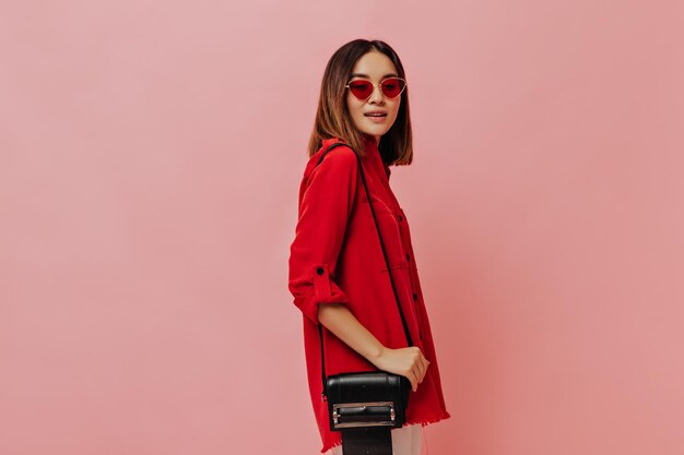 Retrato de uma menina asiática alegre em óculos de sol vermelhos Uma linda mulher de camisa grande segura bolsa preta em fundo rosa