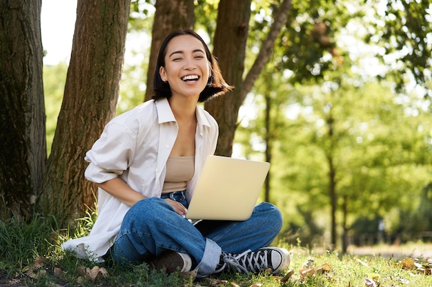 Retrato de uma menina asiática com laptop sentado perto de uma árvore no parque em um dia ensolarado de verão trabalhando em e remoto