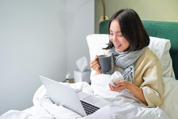 Retrato de uma menina asiática doente deitada na cama com resfriado, com gripe, conversando com o bate-papo por vídeo do laptop trabalhando f