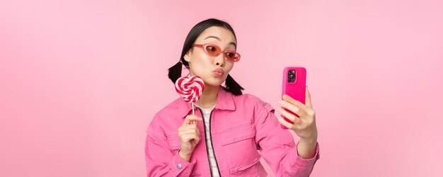 Retrato de uma menina asiática feliz e estilosa tomando selfie com doces de pirulito e sorrindo tirando foto