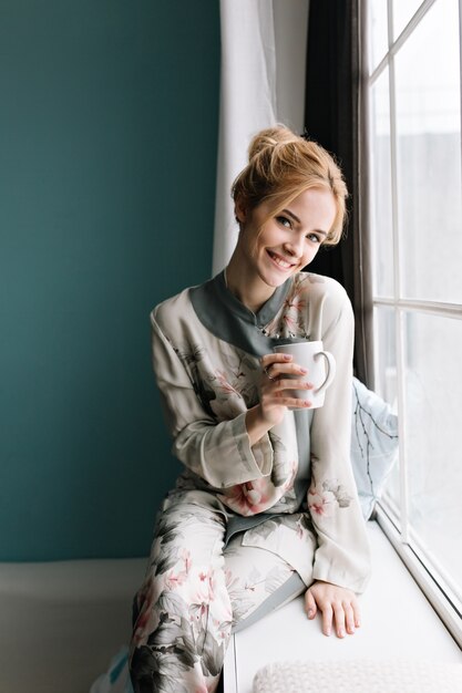 Retrato de uma menina bonita com cabelo loiro preso, sentado no parapeito da janela com uma xícara de café ou chá na mão, tempo de manhã feliz. Parede turquesa. Vestido com pijama de seda com flores.