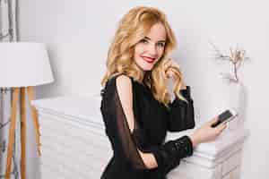 Foto grátis retrato de uma menina bonita loira com smartphone nas mãos em um quarto bem iluminado com interior branco moderno e bonito, em pé contra a lareira falsa, olhando. usando um vestido preto glamurus.
