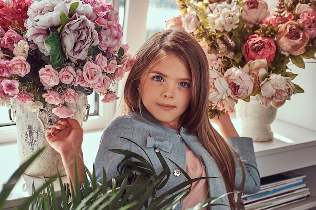 Foto grátis retrato de uma menina bonitinha com longos cabelos castanhos e olhar penetrante, vestindo um vestido elegante, posando com flores contra a janela em casa.