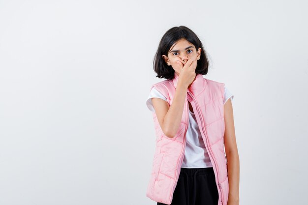 Retrato de uma menina com camiseta branca e colete bufê cobrindo a boca