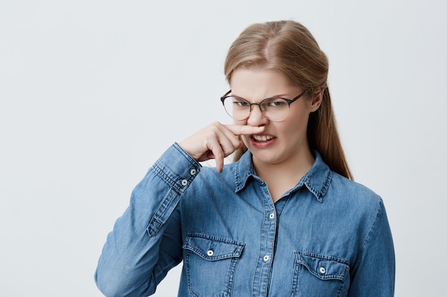 Retrato de uma menina com nojo beliscar o nariz. mulher loira segurando o nariz cheirando algo fedorento. garota estudante vestindo óculos e camisa azul, olhando com nojo. expressão e reação do rosto.