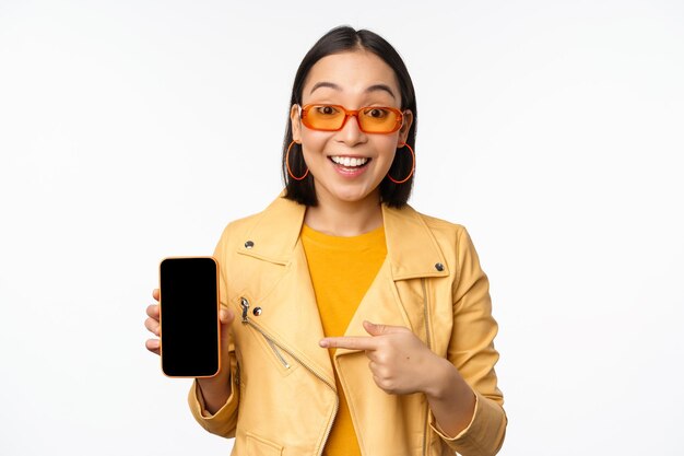 Retrato de uma menina coreana estilosa em óculos de sol sorrindo apontando o dedo para a tela do smartphone mostrando o aplicativo de celular sobre fundo branco