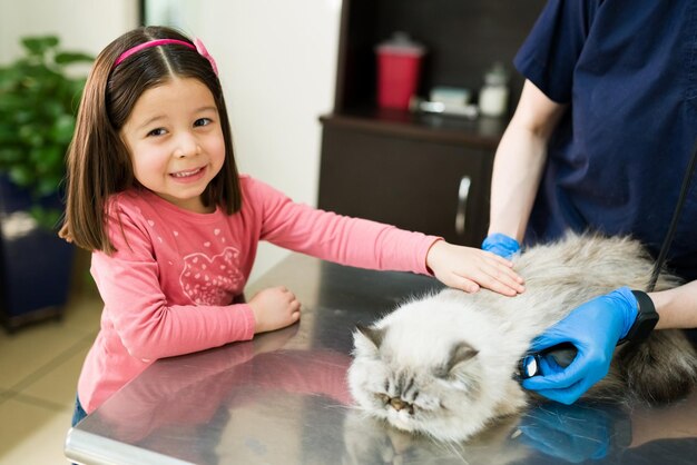 Retrato de uma menina feliz acariciando um gato persa bonito deitado em uma mesa na clínica de animais. Garota elementar levando seu gato branco fofo ao veterinário