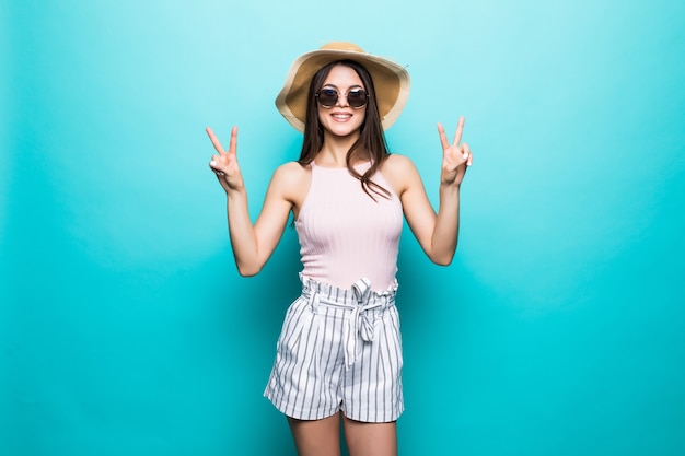 Retrato de uma menina feliz e alegre com chapéu de verão, mostrando um gesto de paz com as duas mãos isoladas sobre uma parede azul.