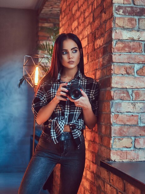 Retrato de uma menina morena atraente, vestida com uma camisa de flanela e jeans com câmera encostada na parede em uma sala com interior loft.