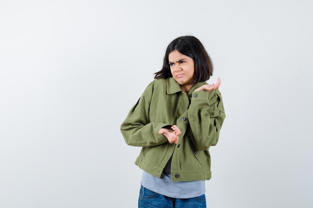 Retrato de uma menina mostrando um gesto de impotência em um casaco, camiseta, jeans e uma vista frontal hesitante