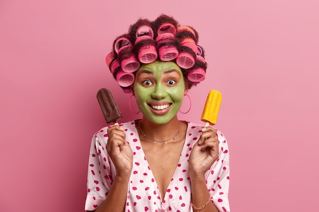 Foto grátis retrato de uma modelo feminina bonita aplica máscara de beleza verde no rosto, usa modeladores de cabelo para fazer cachos, segura sorvete de chocolate e manga, tem bom humor, sorri amplamente, isolado no rosa