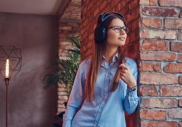 Retrato de uma morena atraente e encantadora de óculos e camisa azul, ouvindo música em fones de ouvido, encostado em uma parede de tijolos em uma sala com design loft.