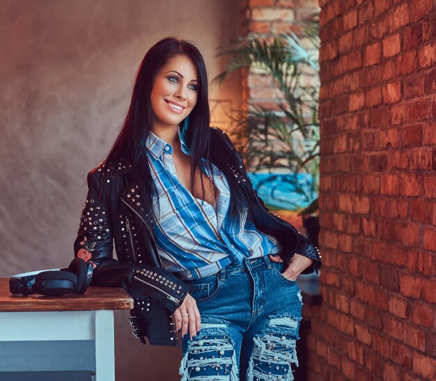 Retrato de uma morena sensual sorridente posando em uma jaqueta de couro elegante e jeans, apoiando-se em uma mesa em um estúdio com um interior loft.