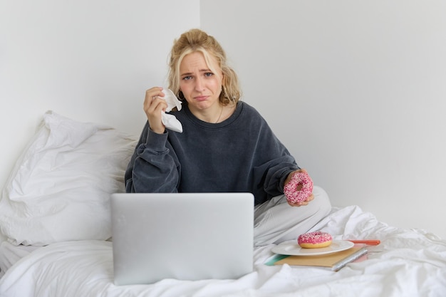 Foto grátis retrato de uma mulher a ver um filme triste no portátil a chorar e a limpar as lágrimas a comer um donut e