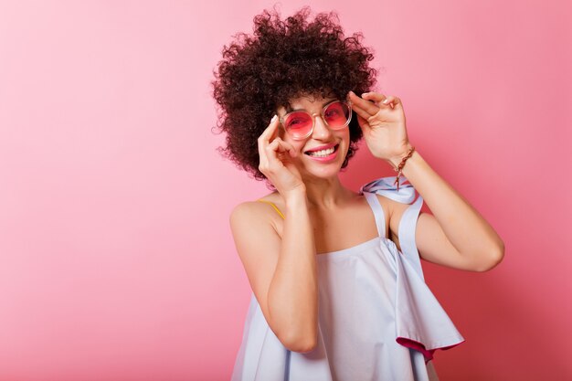 Retrato de uma mulher adorável e ensolarada com cabelo curto e encaracolado e sorriso encantador, usando camisa azul e óculos rosa poses em rosa