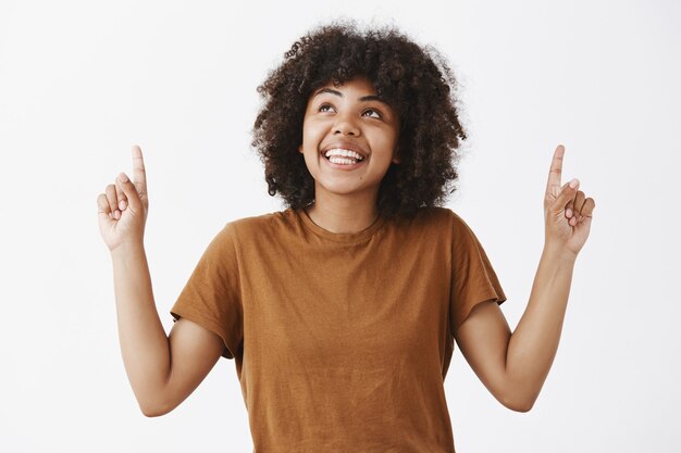 Retrato de uma mulher afro-americana atraente e sonhadora em uma camiseta marrom com penteado afro olhando e apontando para cima com um sorriso alegre