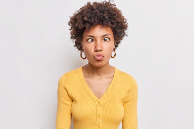 Retrato de uma mulher afro-americana bonita de cabelo encaracolado engraçada mantém os lábios dobrados, cruza os olhos, faz careta na frente, brinca usando um macacão amarelo isolado sobre a parede branca