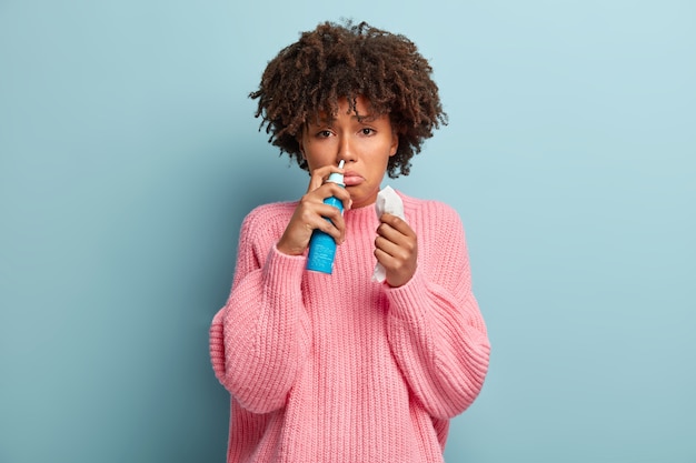 Retrato de uma mulher afro-americana descontente cheira aerossol nasal, sente-se doente, está corando, usa remédio para nariz entupido, segura o lenço, tem expressão facial triste, usa macacão rosa