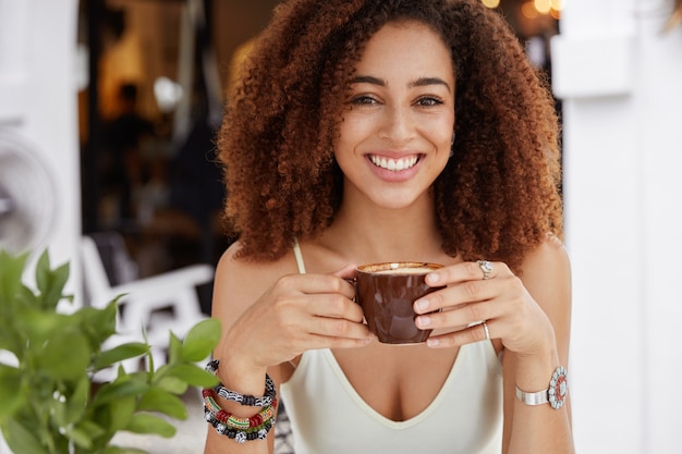 Retrato de uma mulher alegre de pele escura com penteado afro espesso, segurando uma xícara de café ou latte, sentada no interior da cafeteria, de bom humor