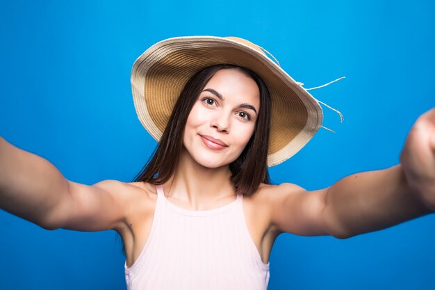 Retrato de uma mulher alegre no vestido e chapéu de verão tomando uma selfie isolada sobre a parede azul.