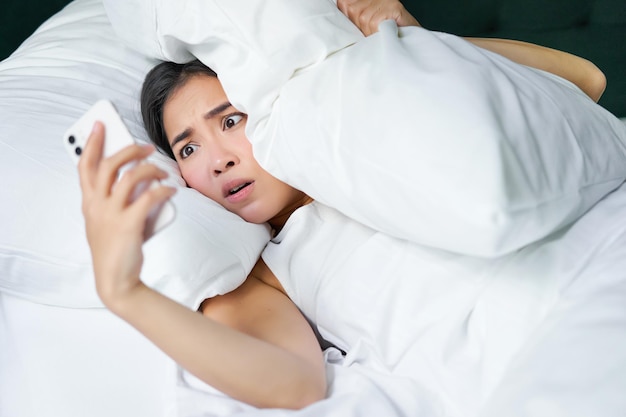 Foto grátis retrato de uma mulher asiática acordando na cama olhando chocada com o celular percebendo que ela dormiu demais