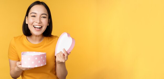 Retrato de uma mulher asiática animada abre a caixa de presentes com uma cara feliz surpresa sobre fundo amarelo