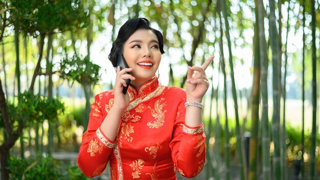 Retrato de uma mulher asiática bonita em um cheongsam chinês sorrindo e apontando o dedo enquanto fala com o smartphone na floresta de bambu