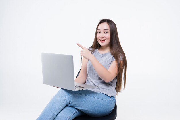 Retrato de uma mulher asiática casual feliz apontando para o lado segurando um laptop enquanto está sentado em uma cadeira sobre uma parede branca
