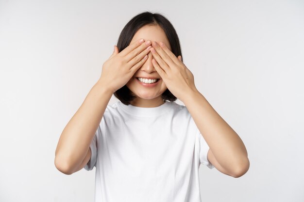 Retrato de uma mulher asiática cobrindo os olhos esperando por surpresa com os olhos vendados sorrindo feliz antecipando a posição contra o fundo branco