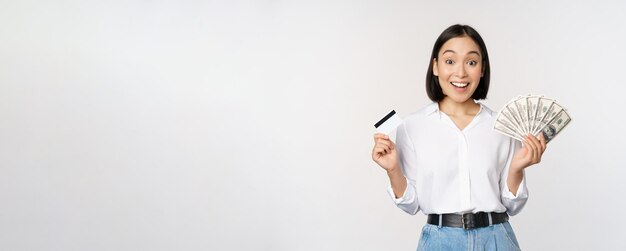 Retrato de uma mulher asiática entusiasmada segurando dinheiro em dinheiro e cartão de crédito sorrindo espantado com o fundo branco da câmera