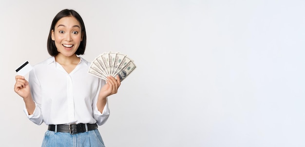 Retrato de uma mulher asiática entusiasmada segurando dinheiro em dinheiro e cartão de crédito sorrindo espantado com o fundo branco da câmera