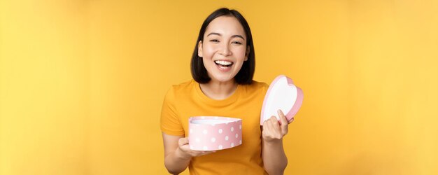 Retrato de uma mulher asiática excitada abre a caixa de presentes com um rosto feliz e surpreso em pé sobre fundo amarelo