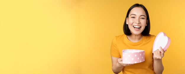 Retrato de uma mulher asiática excitada abre a caixa de presentes com um rosto feliz e surpreso em pé sobre fundo amarelo