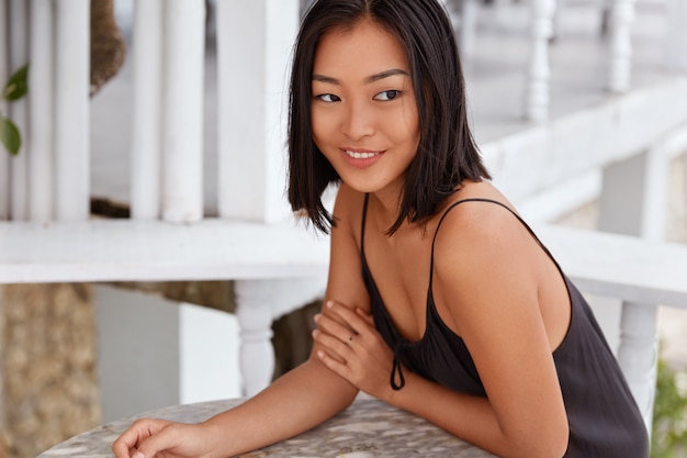 Retrato de uma mulher asiática morena sonhadora com expressão feliz, pensa em algo, vestida de camiseta preta, senta-se contra o interior da cafeteria aconchegante. Pessoas, expressões faciais e conceito de estilo de vida