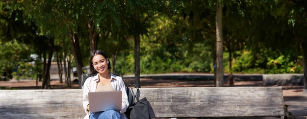 Foto grátis retrato de uma mulher asiática sentada com o laptop no banco do parque ouvindo música com wireless