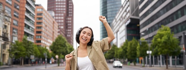 Foto grátis retrato de uma mulher asiática sorridente dançando triunfante, sentindo-se feliz enquanto ouve música na cidade