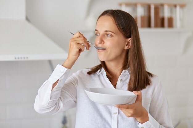 Retrato de uma mulher atraente com fome, com cabelo escuro, camisa branca, olhando para longe, posando na cozinha, tomando café da manhã, segurando o prato e a colher nas mãos.