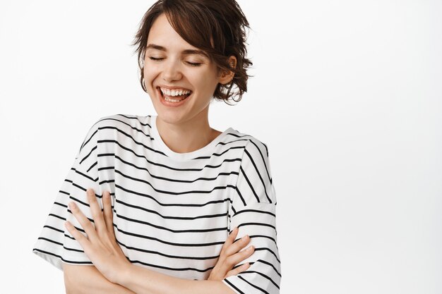 Retrato de uma mulher atraente feliz rindo, braços cruzados no peito, sorrindo com os olhos fechados, em pé em uma camiseta listrada em branco.