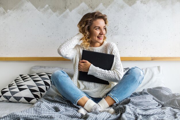 Retrato de uma mulher atraente sentada na cama, com cabelo loiro encaracolado, segurando um livro e um café na xícara.