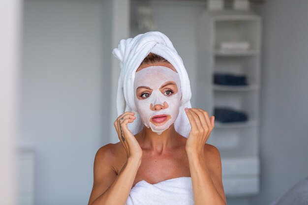 Retrato de uma mulher bonita caucasiana calma com uma toalha na cabeça e uma máscara cosmética no rosto Conceito de cuidados com a pele do rosto Feminino relaxe na cama em casa