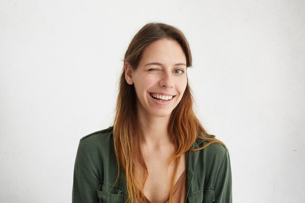 Retrato de uma mulher bonita com cabelo comprido piscando enquanto tem bom humor, sorrindo mostrando seus dentes brancos perfeitos.