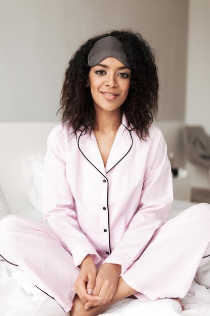 Retrato de uma mulher bonita com cabelo encaracolado escuro, sentado na cama com máscara de olho na cabeça e olhando alegremente na câmera