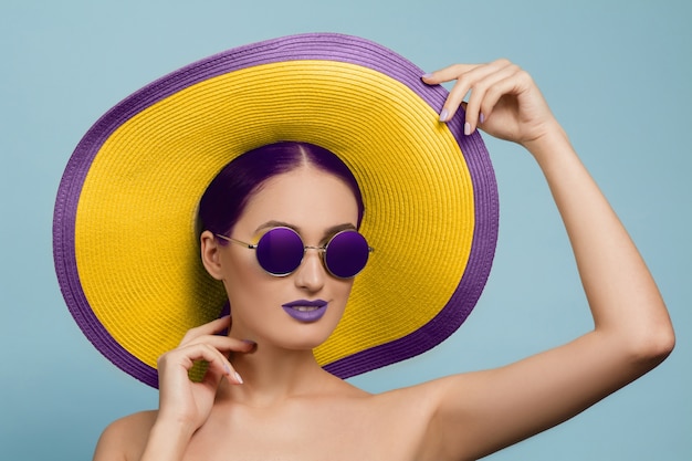 Retrato de uma mulher bonita com maquiagem brilhante, chapéu e óculos de sol no estúdio azul