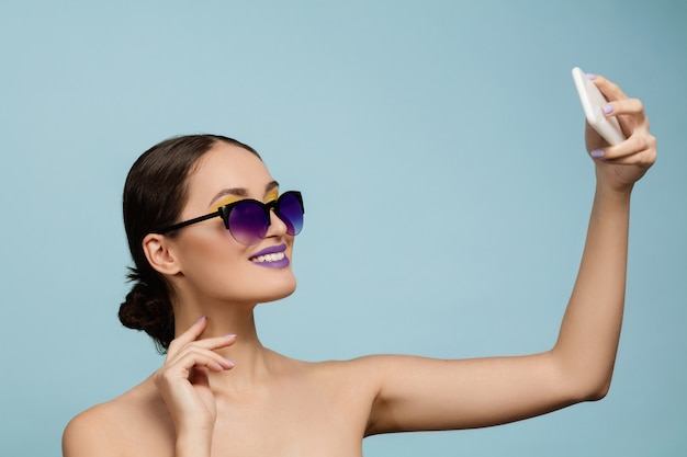 Retrato de uma mulher bonita com maquiagem brilhante e óculos de sol sobre fundo azul do estúdio. Estilo e penteado elegante e moderno. Cores do verão. Conceito de beleza, moda e publicidade. Fazendo selfie.