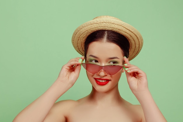 Foto grátis retrato de uma mulher bonita com maquiagem brilhante, óculos vermelhos e chapéu no estúdio verde