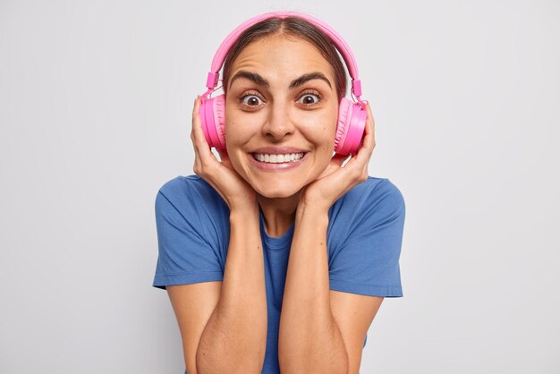 Retrato de uma mulher bonita e alegre usa fones de ouvido rosa sem fio nas orelhas e sorrisos de faixa de áudio com os dentes arreganhados, vestida com uma camiseta azul casual isolada sobre a parede branca