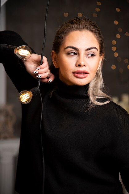 Retrato de uma mulher bonita feliz em uma camisola escura em pé perto de lâmpadas.