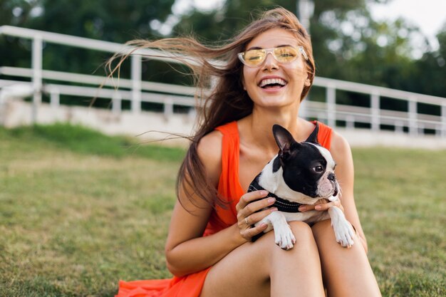 Retrato de uma mulher bonita feliz sentada na grama no parque de verão, segurando o cachorro Boston Terrier, sorrindo, humor positivo, brincando com o animal de estimação, se divertindo