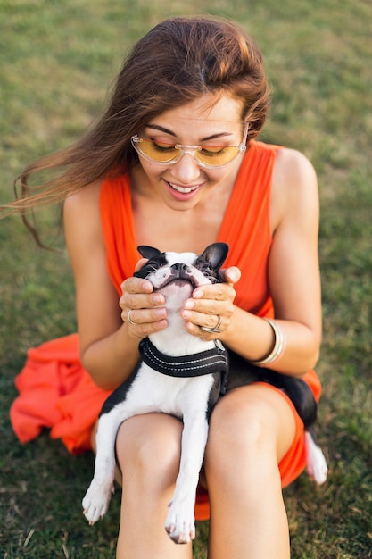 Retrato de uma mulher bonita feliz sentada na grama no parque de verão, segurando o cachorro Boston Terrier, sorrindo, humor positivo, vestindo um vestido laranja, estilo moderno, óculos de sol, brincando com o animal de estimação